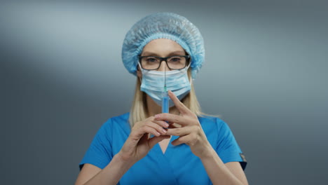 Close-up-of-the-young-blonde-Caucasian-woman-doctor-in-hat,-glasses-and-mouth-mask-holding-a-syringe-with-a-needle.
